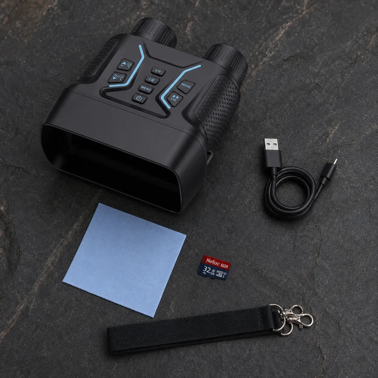 A black all-in-one 4K infrared night vision recording binocular with buttons, a USB cable, a cleaning cloth, a memory card, and a strap on a dark stone surface.